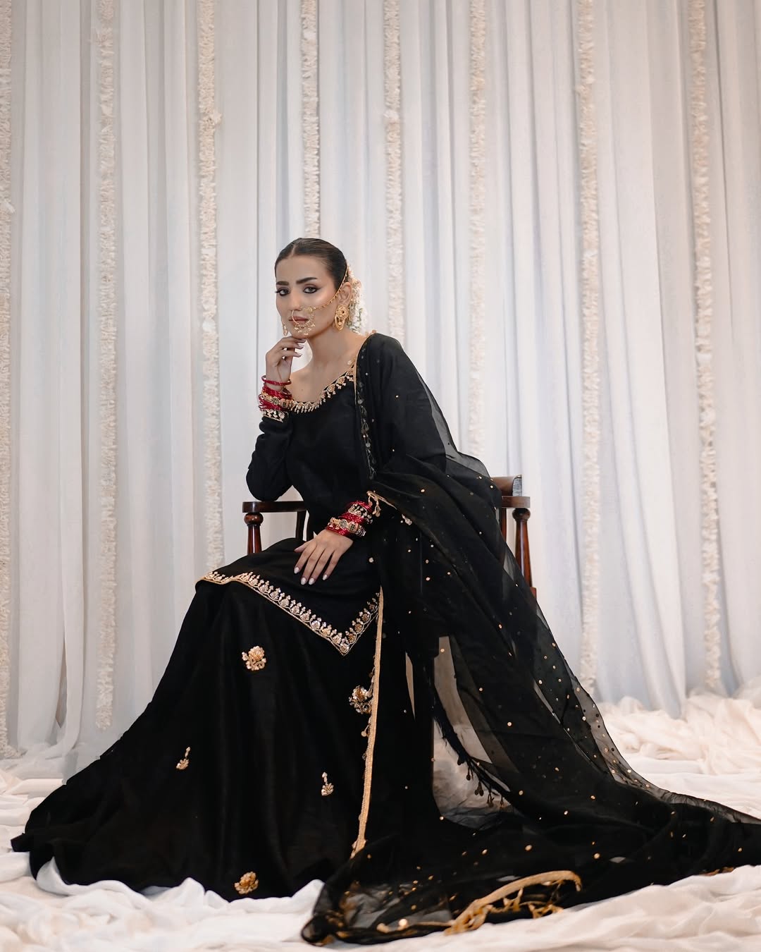 A woman seated on a wooden chair wearing a black traditional outfit with gold embroidery and a sheer black dupatta, posed against a white draped backdrop with vertical gold decorative accents.