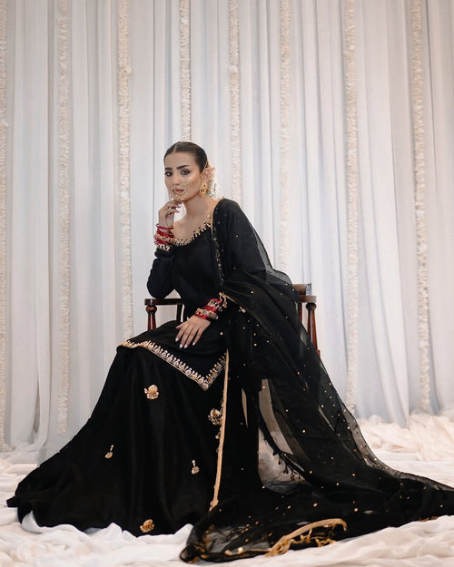A woman seated on a wooden chair wearing a black traditional outfit with gold embroidery and a sheer black dupatta, posed against a white draped backdrop with vertical gold decorative accents.