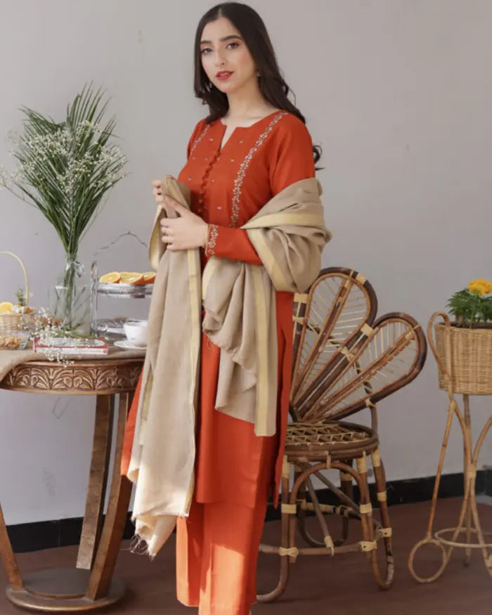 Woman in an orange traditional outfit holding a beige scarf in a room with a table and chairs.